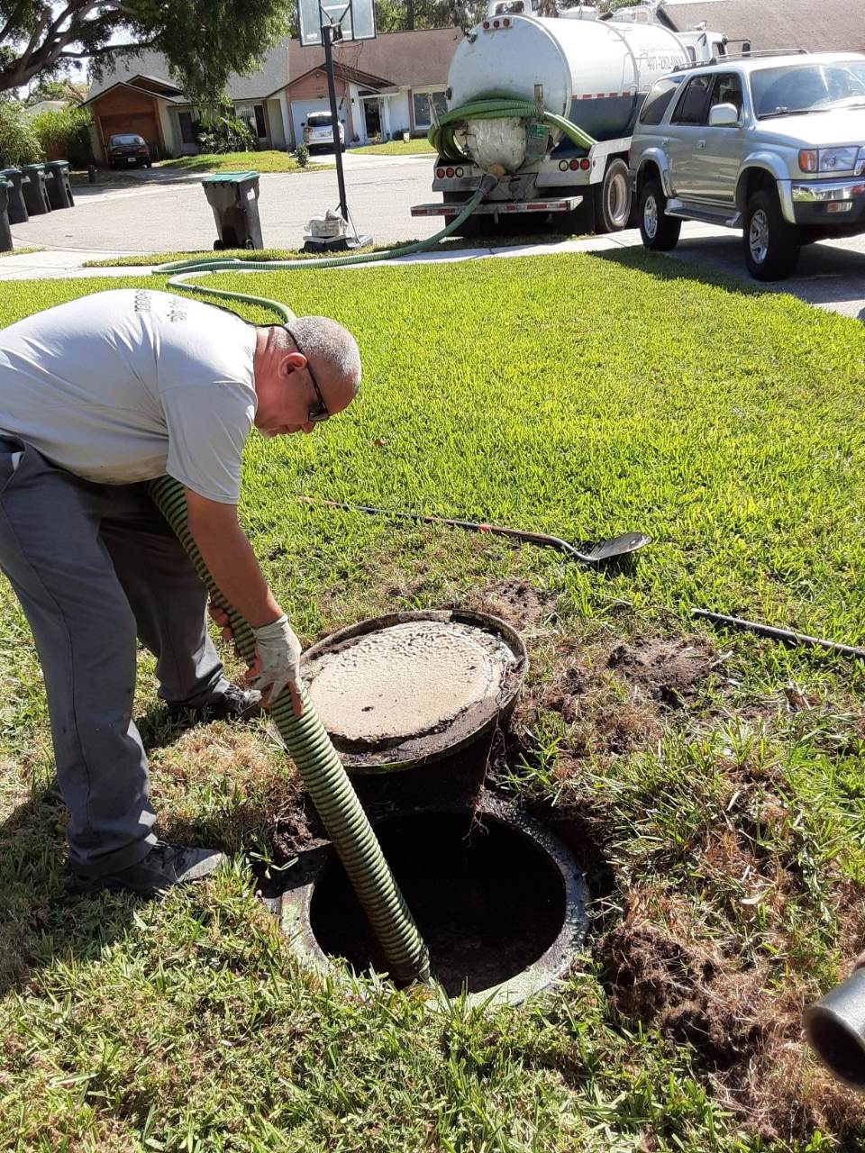 Worker in a residential yard uses a hose to pump sewage from an open drain, with a tanker truck and pickup parked behind him.