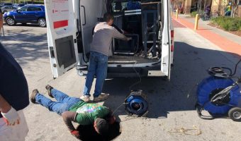 A person lies on the pavement beside an open service van while another assists; mobility scooters and onlookers surround the tense scene.