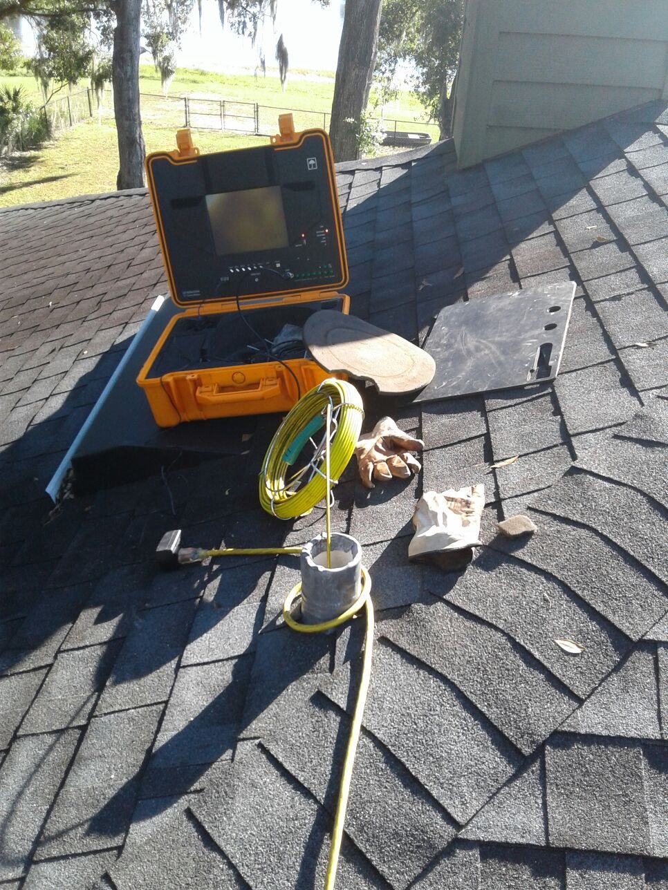 Open orange repair case and tools sit on a shingled rooftop beside a yellow hose, with bright sunlight, trees, and a house in the background.