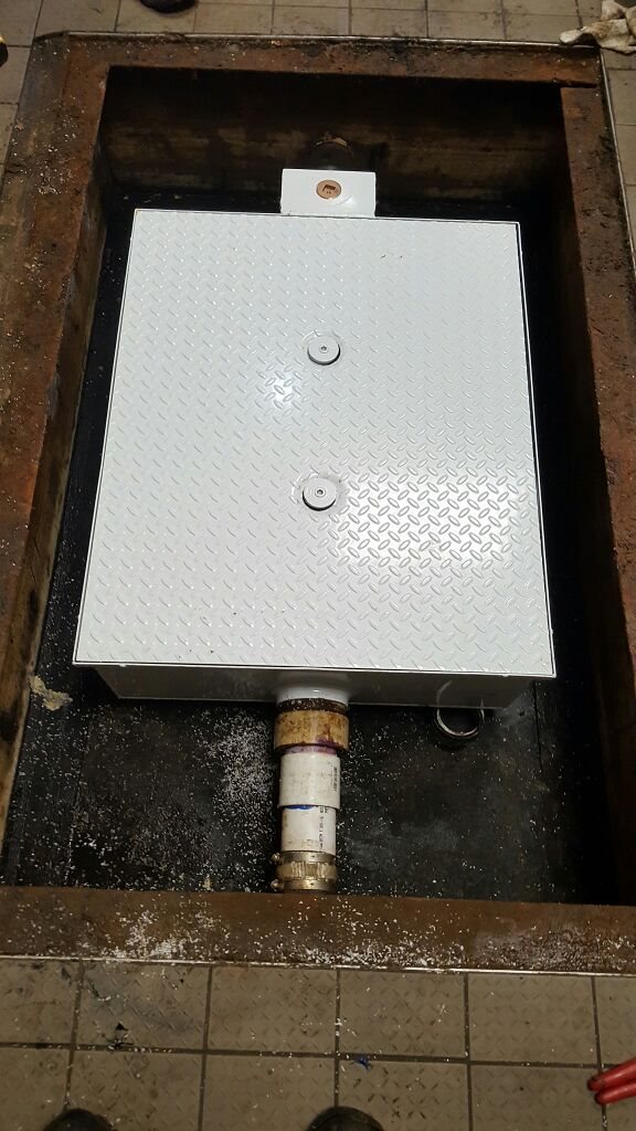 White diamond-plate metal access panel with two circular fasteners in a rusty floor opening, connected to a pipe; worn industrial setting.