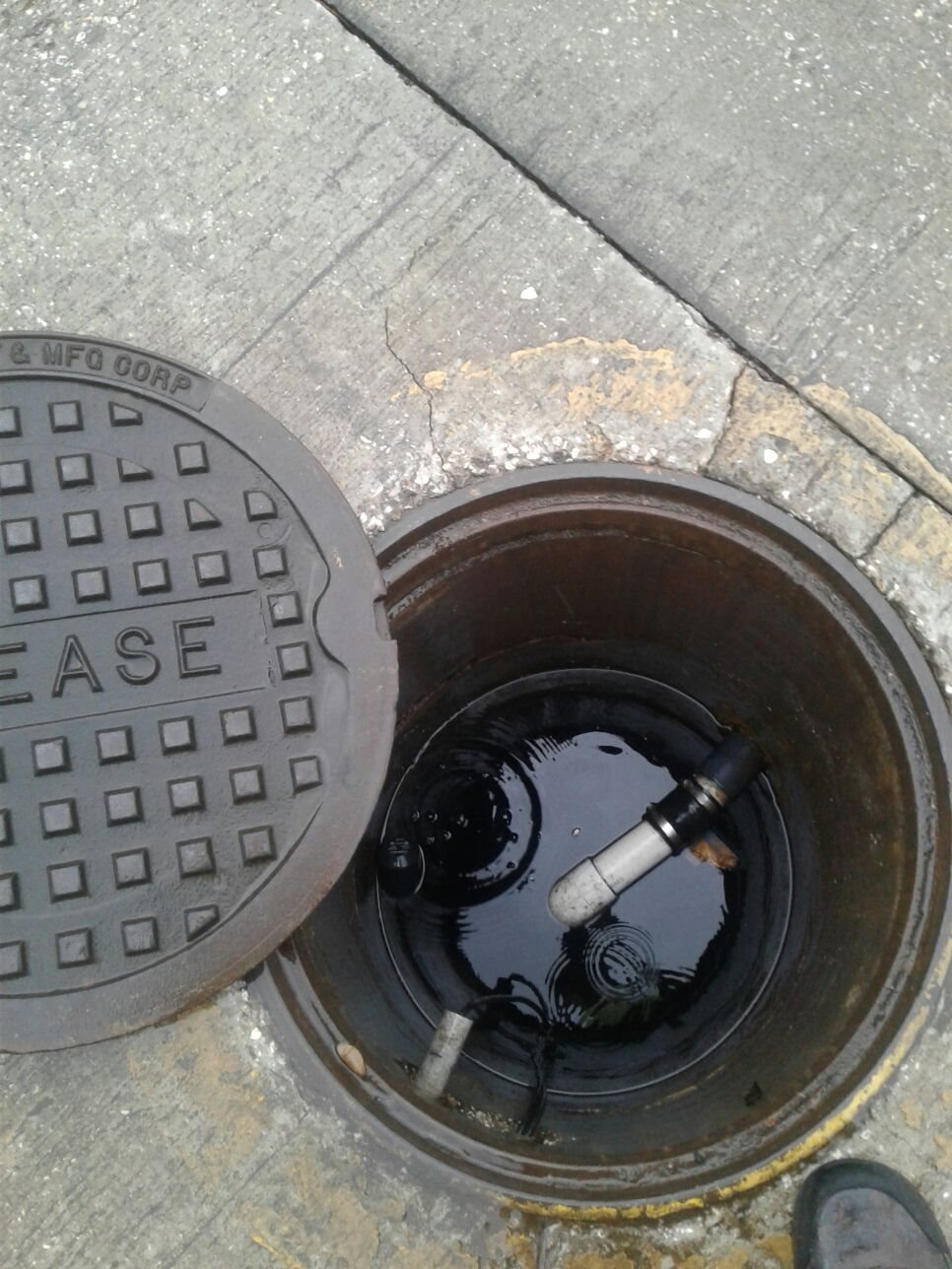 Open manhole beside its patterned cover on a concrete sidewalk, filled with dark rainwater and a submerged pipe; somber urban scene.